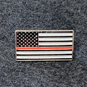 May include: A small, metal pin featuring a red, white, and blue American flag with a single red stripe across the middle. The flag is on a dark gray background.