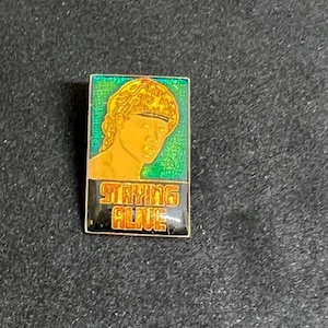 May include: A gold and black enamel pin with a green background. The pin features a cartoon image of a person with blonde hair and the text "Staying Alive".