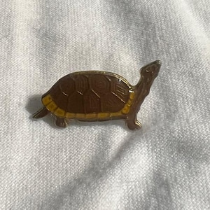 May include: A small, brown turtle pin with a yellow shell. The pin is made of metal and has a detailed design.