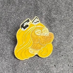 May include: A yellow and black enamel pin featuring a cartoon character with long blonde hair and a brown face. The character is wearing a black bow.