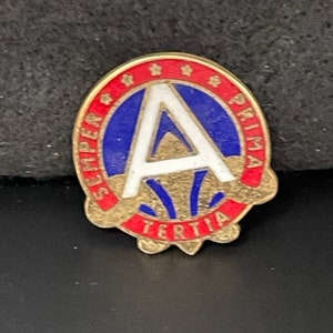 May include: A round, gold-toned pin with a red and blue design. The pin features a white letter "A" in the center, surrounded by text and stars. The words "Semper Prima Tertia" are visible.