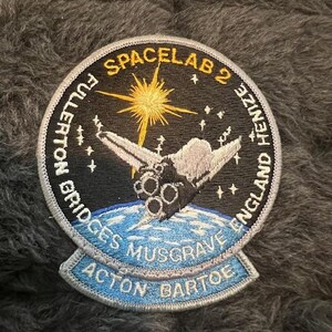 May include: A blue and white embroidered patch with the text "SPACELAB 2 FULTON BRIDGES MUSGRAVE ENGLAND HENIZE ACTON BARTOE" around a black background with a white space shuttle and a yellow star.