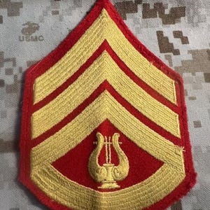May include: A red and gold military rank insignia with three gold stripes and a gold lyre symbol. The insignia is for a Sergeant.