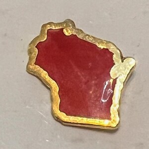 May include: A gold-toned pin in the shape of the state of Wisconsin. The interior is a deep red color, and the edges are outlined with a textured gold. The pin is a small, decorative item, likely a souvenir or a piece of jewelry.