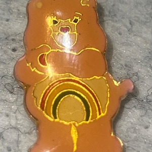 May include: A gold-toned metal enamel pin shaped like a bear with light brown fur. The bear features a central belly graphic depicting a colorful rainbow with red, yellow, and green arches. The pin has a glossy finish and gold-colored metal outlines.