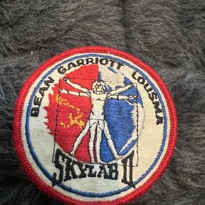 May include: A white, red, and blue embroidered patch with the text "BEAN GARRIOTT LOUSMA SKYLAB II". The patch features a stylized image of a person with outstretched arms.