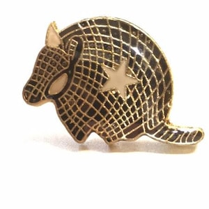 May include: A gold-tone pin featuring a black and white armadillo with a white star on its back.