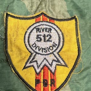 May include: A yellow shield-shaped patch with black trim. The patch features a white circle with the text "RIVER 512 DIVISION" and the letters "PBR" in red. The background is camouflage fabric.