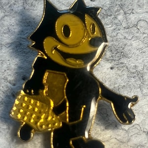 May include: A black and gold enamel pin featuring the cartoon character Felix the Cat. The pin depicts Felix with his signature grin, holding a bag. The pin has a gold border and accents, and is set against a gray background.