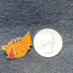 May include: A gold-colored pin of a yellow trumpet with red and black details. The pin is next to a US quarter for size comparison.