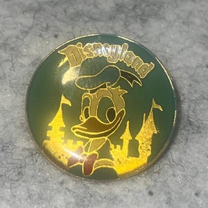 May include: A round metal pin featuring a green background with a gold-toned illustration of Donald Duck and a castle silhouette. The word Disneyland is printed in gold lettering at the top of the pin, which has a metallic gold-colored border.