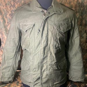 May include: Olive green military-style jacket with multiple pockets and a zip-up front. The jacket has a collared neckline and a drawstring closure. The jacket is displayed on a mannequin. The jacket is made of a durable, possibly water-resistant material.