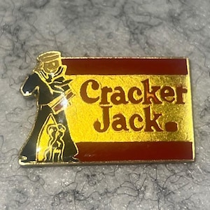 May include: A rectangular pin featuring the Cracker Jack logo. The pin has a gold background with the words "Cracker Jack" in red lettering. A sailor figure in black and gold is on the left side of the pin.