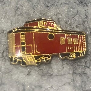 May include: A red and gold enamel pin in the shape of a caboose. The pin features a detailed design with black accents, including wheels and windows. The caboose has a gold outline and is set against a textured background.