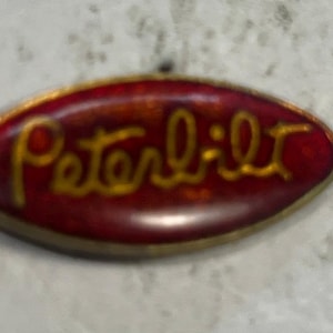 May include: Oval-shaped Peterbilt emblem with a red background and gold lettering. The emblem has a gold border and is slightly blurred. The Peterbilt logo is a classic design, often seen on trucks.