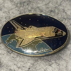 May include: An oval-shaped enamel pin featuring a space shuttle flying above a blue and green Earth against a dark blue starry sky. The pin has a gold-toned metal border and depicts the shuttle in shades of white, gray, and black.