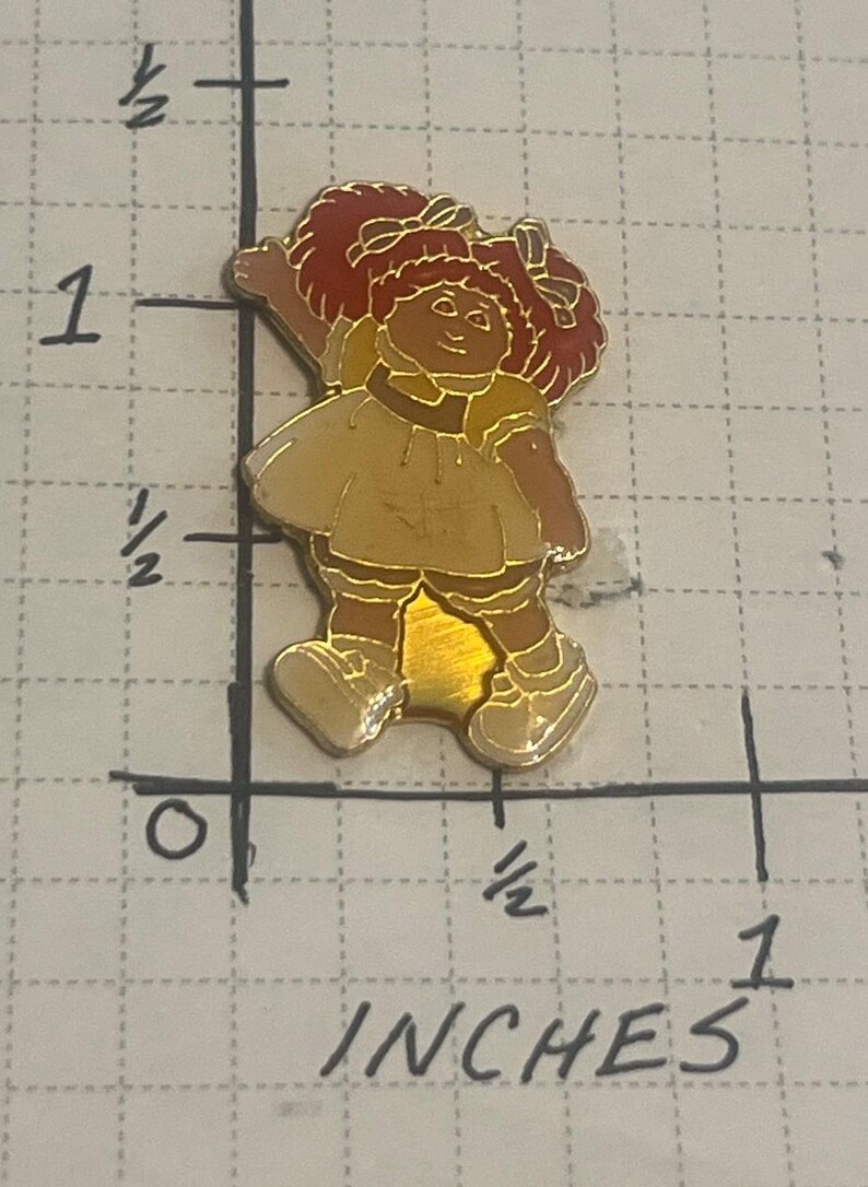 May include: A gold-toned metal enamel pin featuring a character with red pigtails, a yellow dress, and white shoes. The pin is positioned on a grid marked in inches, showing the figure measures approximately 1.25 inches in total height.