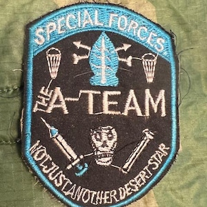 May include: A black and blue embroidered patch with the text "Special Forces The A-Team Not Just Another Desert Star". The patch features a blue arrowhead with three arrows pointing out from it, a skull and crossbones, and two syringes.