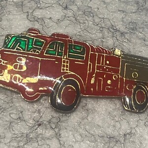 May include: A red and gold-toned fire truck pin with green window detailing. The pin features black wheels and gold trim, showcasing a classic fire engine design. This collectible pin is a great gift for firefighters or fire truck enthusiasts.