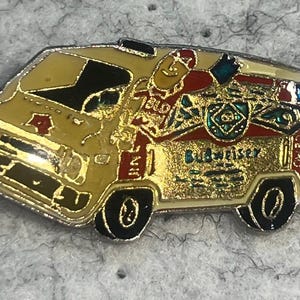 May include: A gold-colored pin in the shape of a van, featuring a cartoon figure and the Budweiser logo. The van has black wheels and windows, with red and blue accents. The text "Budweiser" is visible on the side.