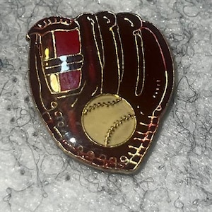 May include: A baseball-themed pin featuring a brown baseball glove and a baseball. The glove has gold trim and a red accent. The baseball is cream-colored with detailed stitching.