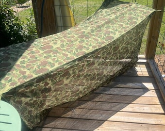Op de afbeelding: Een camouflagezeil, met groene, bruine en beige patronen, is over een houten constructie gedrapeerd. Het zeil is gedeeltelijk opgehangen, waardoor een beschutte ruimte ontstaat. Het houten dek en een groene tafel zijn zichtbaar.
