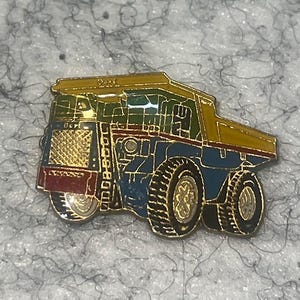 May include: A colorful enamel pin depicting a large dump truck. The pin features a yellow cab, blue body, and black tires with gold accents. The design includes red, green, and gold details, creating a vibrant and detailed miniature.