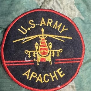 May include: A black and red circular patch with the words "U.S. ARMY" in yellow lettering above a yellow helicopter with red accents. The words "APACHE" are in yellow lettering below the helicopter.