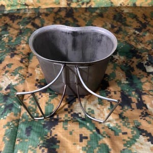 May include: A silver metal canteen with a folding handle. The canteen is on a camouflage fabric background.