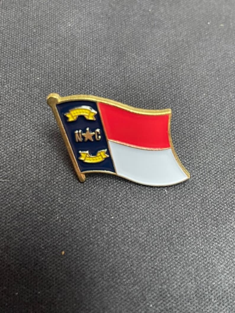 May include: A gold-colored pin with the North Carolina state flag. The flag is red, white, and blue with a yellow star in the blue section. The words "N.C." are printed in yellow on the blue section.