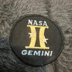 May include: A black and white NASA Gemini patch with a gold Roman numeral II and two white stars. The text "NASA" and "GEMINI" are in white.