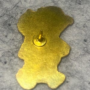 May include: A gold-toned metal pin back featuring a smooth, irregular silhouette that follows the shape of a character. A single centered metal post protrudes from the flat surface, designed for attachment to fabric or other materials.