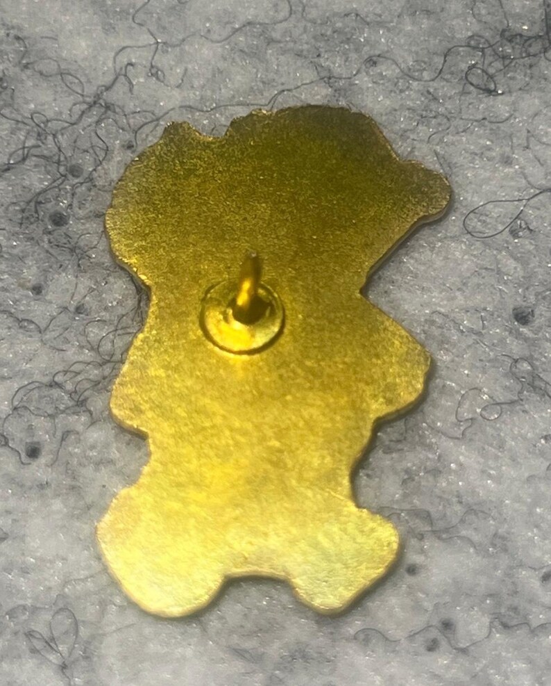 May include: A gold-toned metal pin back featuring a smooth, irregular silhouette that follows the shape of a character. A single centered metal post protrudes from the flat surface, designed for attachment to fabric or other materials.