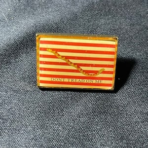 May include: A gold-colored pin with a red and white striped flag featuring a yellow snake with the text "DONT TREAD ON ME".