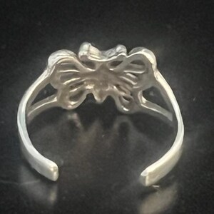 May include: A silver-toned toe ring with a butterfly design. The ring features an open design with a butterfly in the center and curved bands that wrap around the toe. The ring is set against a dark background.