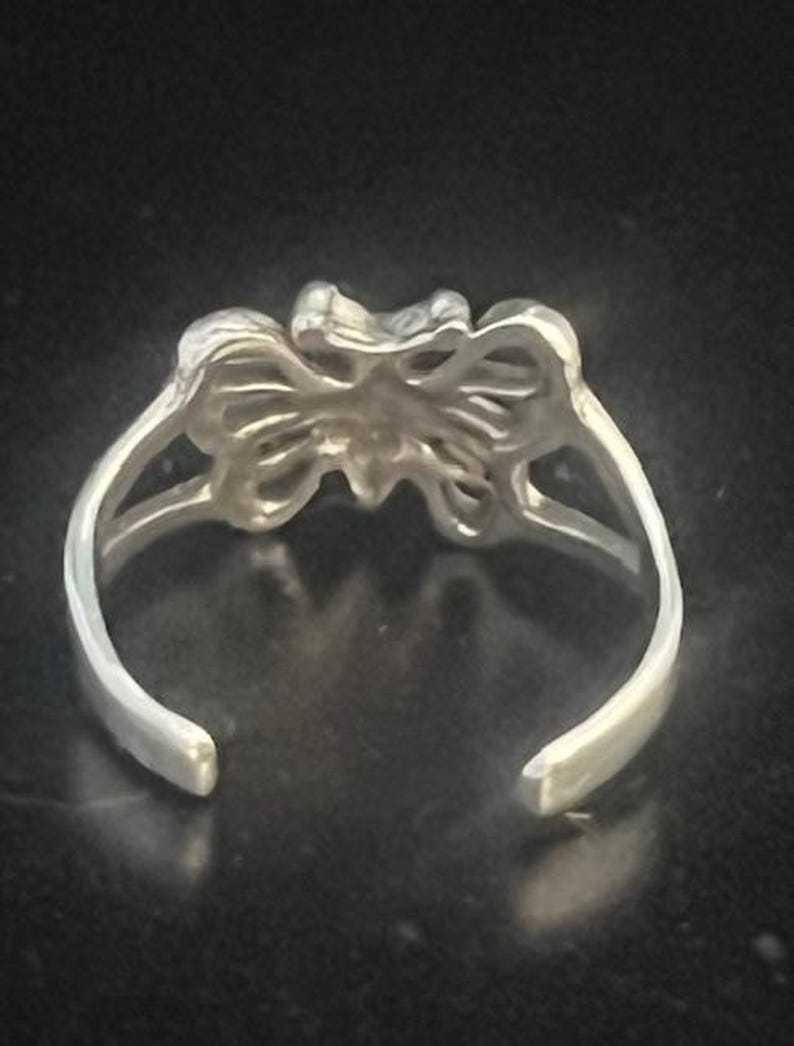 May include: A silver-toned toe ring with a butterfly design. The ring features an open design with a butterfly in the center and curved bands that wrap around the toe. The ring is set against a dark background.