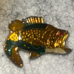 May include: A detailed pin in the shape of a fish, featuring a gold body with green and yellow accents. The fish has a red eye and is set against a textured, gray background. The pin is likely made of metal and enamel.