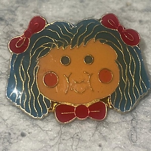 May include: A metal enamel pin shaped like a doll head with wavy dark teal hair, two red hair bows, a tan face, round red cheeks, and a red bow tie at the chin. The design features gold-toned metal outlines separating the colored enamel sections.