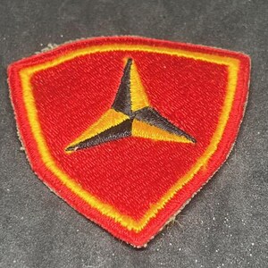 May include: Embroidered patch with a red shield shape, a yellow border, and a black and yellow starburst design in the center. The patch is likely for a uniform or garment.