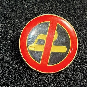 May include: A round, metal pin with a red border and a black center. The center features a yellow cigarette with a red slash through it, indicating a no-smoking symbol. The pin has a gold-colored edge.