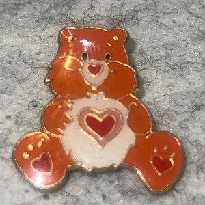 May include: A metal enamel pin shaped like a seated orange bear with a light pink belly patch featuring a dark red heart. The bear has a small red heart on its nose and a red heart on each foot pad, all outlined in a gold-toned metal finish.