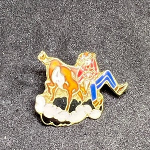 May include: A gold-colored pin with a cartoon image of a cowboy riding a bucking bronco. The cowboy is wearing a red shirt, blue pants, and a brown hat. The bronco is brown with white markings. The pin is set against a black background.