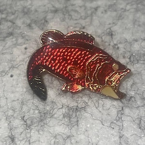 May include: A red and gold fish-shaped pin with a detailed design. The pin features a textured red body with gold accents, including the fins, mouth, and eye. The fish has a black tail and is likely made of metal.