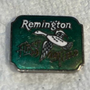 May include: A rectangular, vintage Remington First in the Field pin. The pin features a green background with the word "Remington" in white script at the top. A white and black duck graphic is in the center. The words "First in the Field" are at the bottom.
