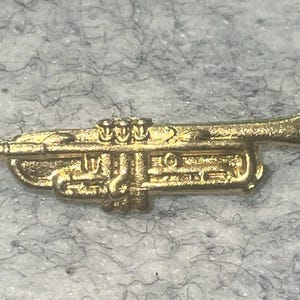 May include: A small, gold-colored trumpet pin. The miniature brass instrument features detailed valves, tubing, and a bell. The pin is likely made of metal and is designed to be worn as an accessory. The trumpet is a musical instrument.