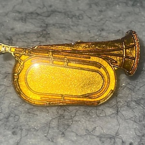 May include: A gold-colored brass instrument pin with a detailed design. The pin features a trumpet shape with a bell, valves, and tubing. The pin is likely made of metal and has a shiny, reflective surface. The pin is a musical instrument accessory.