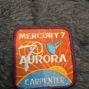 May include: A maroon embroidered patch with an orange background. The patch features a white stylized rocket with the text "MERCURY 7 AURORA CARPENTER" in white.