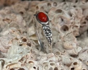 Op de afbeelding: Een zilveren ring met een ovale, helderrode edelsteen. De ring heeft een bloemmotief op de band en is geplaatst tegen een getextureerde, gebroken witte achtergrond. Het is een sieraad.