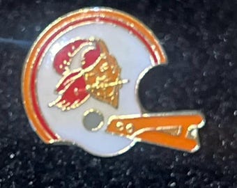 Op de afbeelding: Een emaille pin in de vorm van een American footballhelm in wit, oranje en rood. Het helmdesign heeft een rood en goudkleurig logo. De pin heeft een gouden rand en een speldsluiting.
