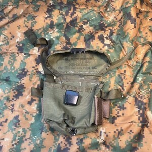 May include: An open, olive drab military ammunition pouch with a black clasp and a small pocket. The pouch has straps and a metal grommet. Text inside the pouch reads: "CASE AMMUNITION M16, 30 ROUND MAGAZINES... EASTERN CANVAS PROD. INC."